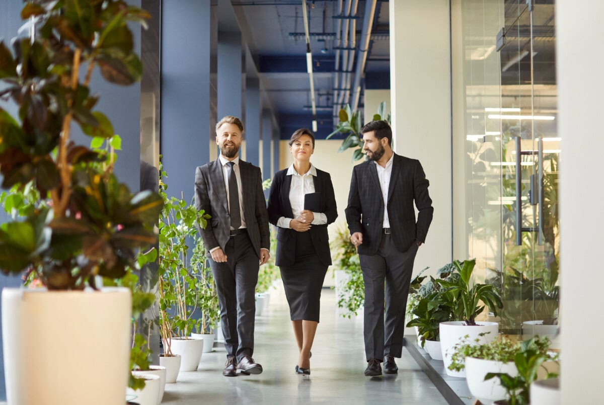 confident business professionals walking through office corridor with green plants, discussing work.
